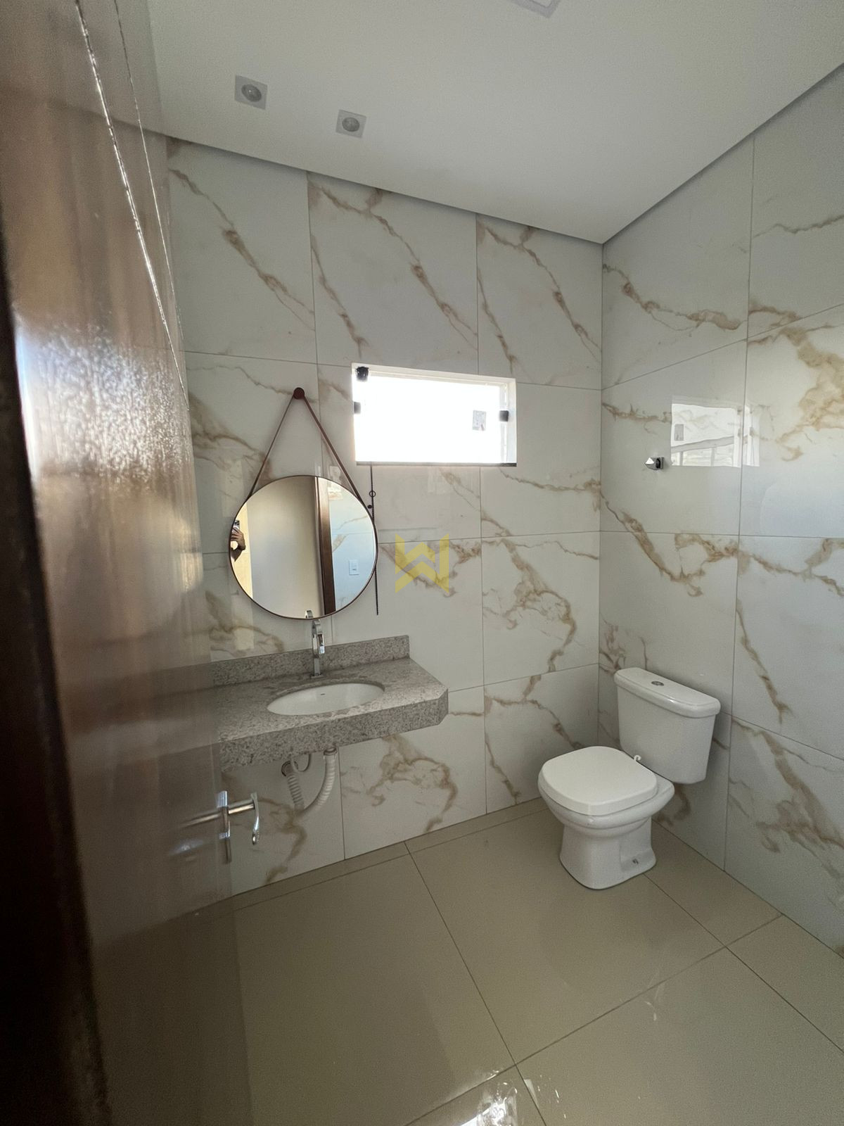IMG-20251021-WA0009 Banheiro moderno em imóvel à venda em Vilhena - RO, com revestimento em porcelanato marmorizado, pia de granito, espelho redondo e vaso sanitário branco.