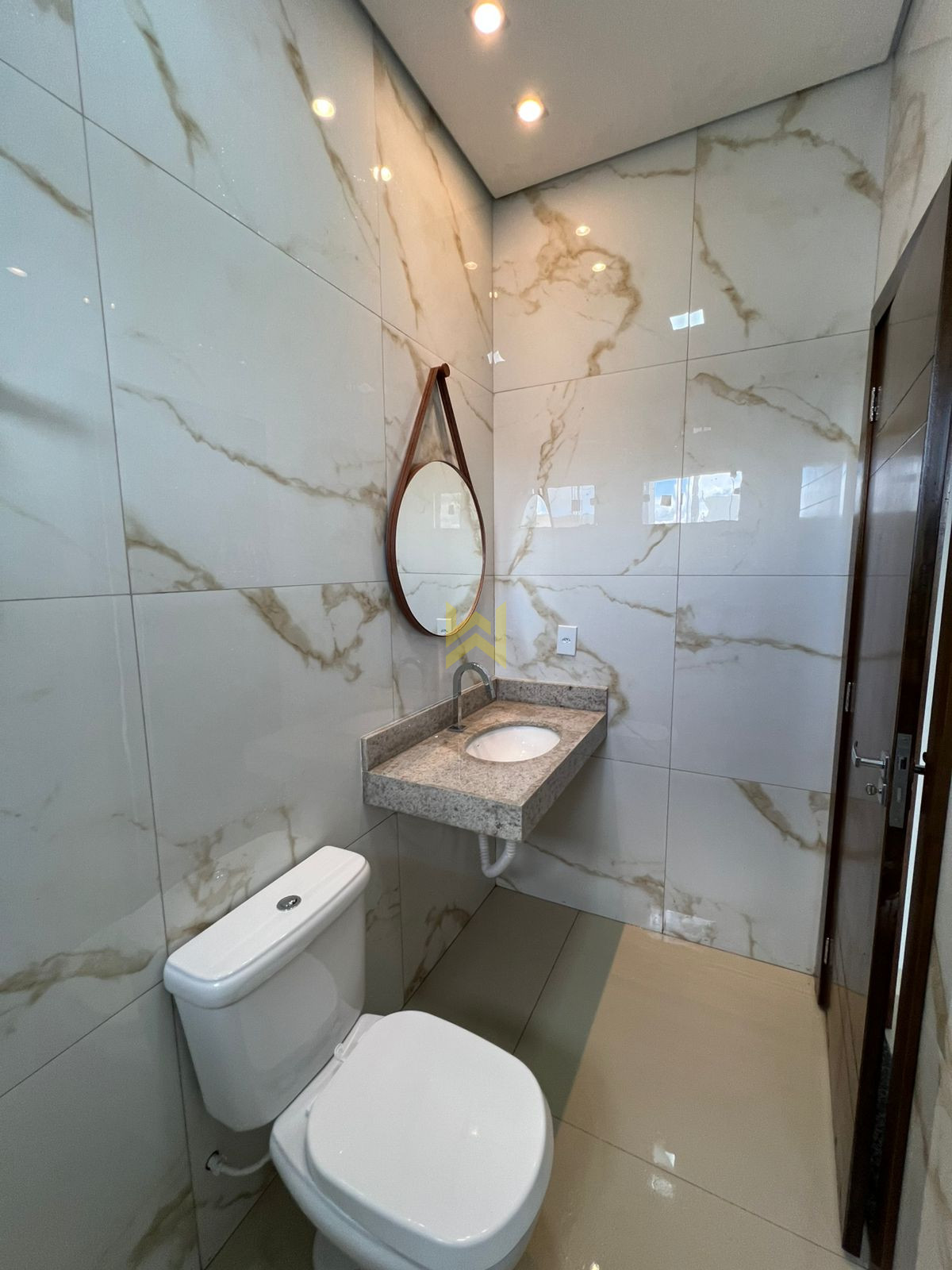 IMG-20251021-WA0011 Banheiro moderno em imóvel à venda em Vilhena - RO, com revestimento em porcelanato marmorizado, pia de granito, espelho redondo e vaso sanitário branco.